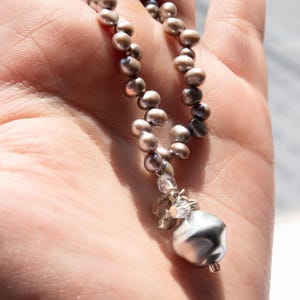 May include: A necklace featuring a strand of small, gray pearls. The necklace has a cluster of clear crystals and a large, irregularly shaped silver pendant. The jewelry is held in a hand, with sunlight illuminating the details.