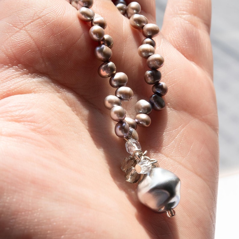 May include: A necklace featuring a strand of small, gray pearls. The necklace has a cluster of clear crystals and a large, irregularly shaped silver pendant. The jewelry is held in a hand, with sunlight illuminating the details.