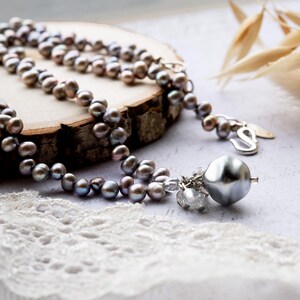 May include: A necklace featuring a strand of gray pearls with a silver clasp and a large, silver pendant. The pearls have a subtle iridescent sheen, and the pendant is a smooth, rounded shape. The necklace is displayed on a wooden surface.
