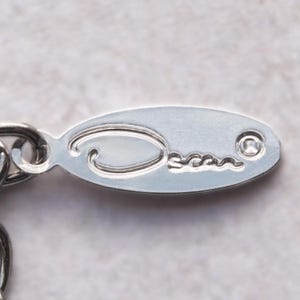 May include: Close-up of a silver oval-shaped tag with the word "Oscar" engraved on it. The tag is attached to a silver chain. The background is a light gray color.