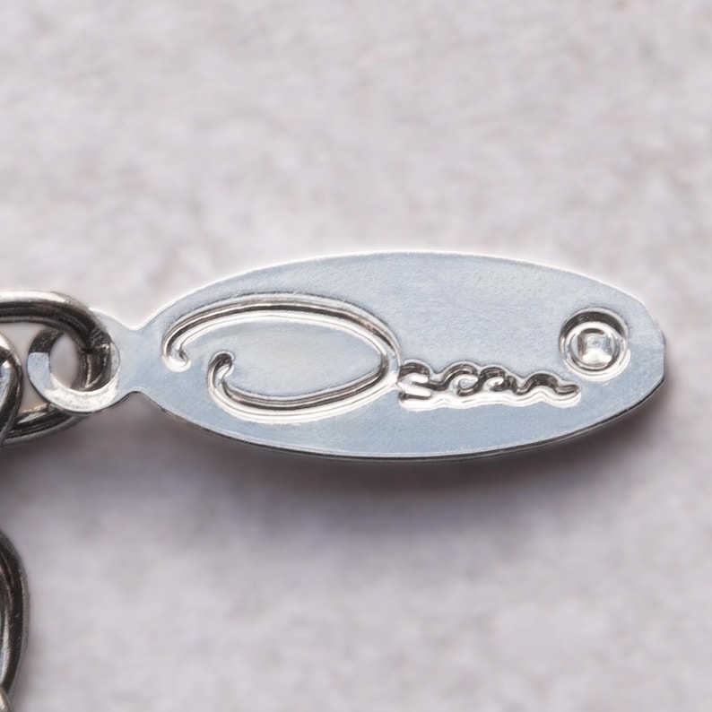 May include: Close-up of a silver oval-shaped tag with the word "Oscar" engraved on it. The tag is attached to a silver chain. The background is a light gray color.