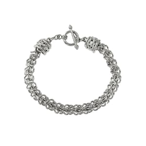 Puede incluir: Una pulsera plateada con cierre de palanca. La pulsera est&aacute; hecha de peque&ntilde;os anillos interconectados, creando un efecto de cota de malla. Cuentas cil&iacute;ndricas decorativas con intrincados patrones calados se encuentran en cada extremo de la pulsera.