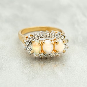 May include: A gold ring with three oval-shaped white opals set in a row. The opals are surrounded by a halo of small clear rhinestones.