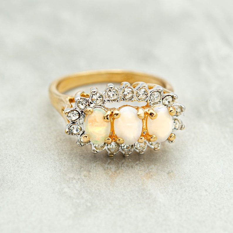May include: A gold ring with three oval-shaped white opals set in a row. The opals are surrounded by a halo of small clear rhinestones.