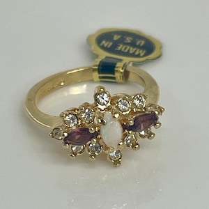 May include: A gold ring with a white opal and purple stones surrounded by clear rhinestones. The ring has a tag that says "Made in U.S.A."