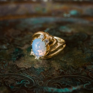 May include: A gold ring featuring a large oval opal gemstone, surrounded by smaller clear gemstones. The ring is displayed on a textured, metallic surface.