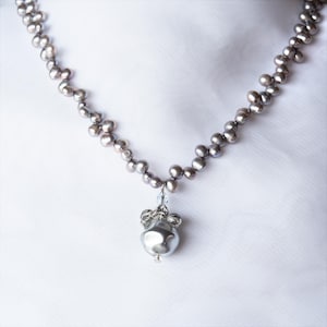 May include: A silver-toned necklace featuring a strand of small, dark gray pearls. The necklace has a pendant with a large, irregularly shaped pearl and a decorative silver element. The background is a white fabric.