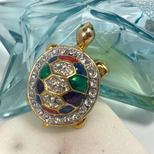 May include: A gold-toned turtle brooch with a colorful, geometric shell design. The shell features red, green, blue, and purple segments, each outlined in gold and encrusted with clear crystals. The turtle's head and feet are also gold, with green accents.