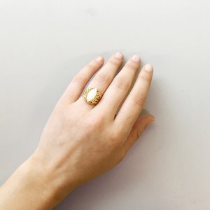 May include: A gold ring with a large white oval-shaped stone set in a detailed, ornate design.
