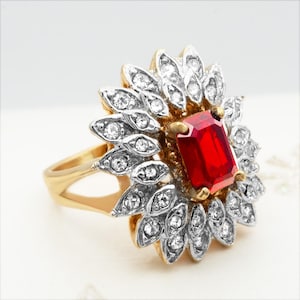 Può includere: Un anello d'oro con una grande gemma rossa rettangolare al centro, circondata da un alone di gemme chiare più piccole. Il design dell'anello include una disposizione floreale delle pietre più piccole, creando un aspetto sorprendente ed elegante.