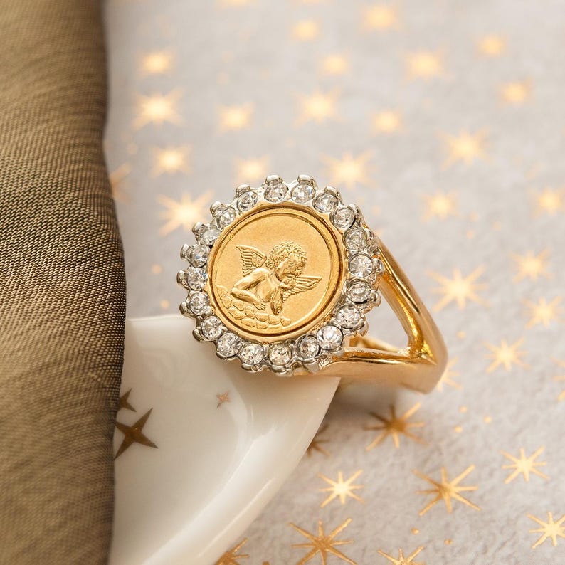 May include: A gold-toned ring featuring a circular design with a detailed angel relief. The ring is encircled by a halo of clear, sparkling gemstones. The ring is set against a background with star-shaped patterns.