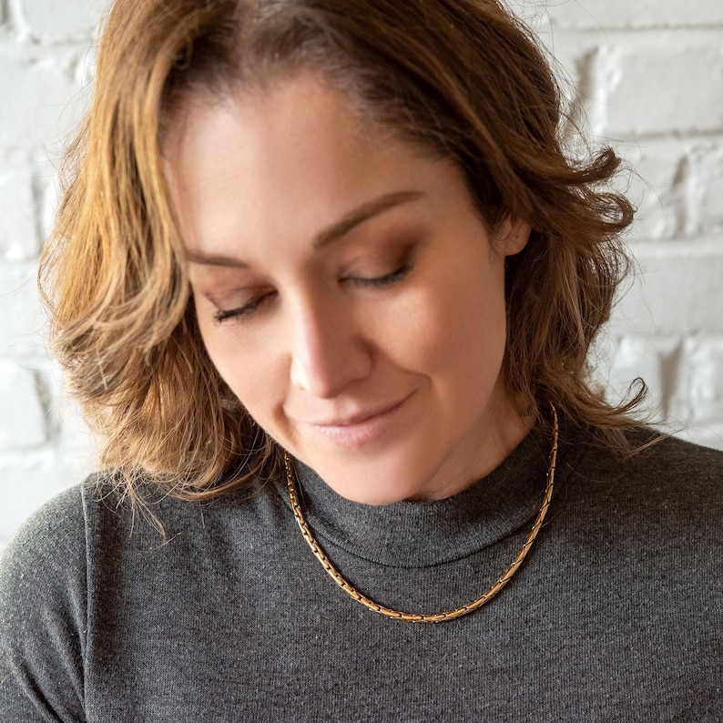 May include: A woman with long brown hair wearing a gray turtleneck sweater with a gold chain necklace.