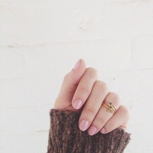 May include: A hand wearing three gold rings with small green stones. The hand is wearing a brown knitted sweater.