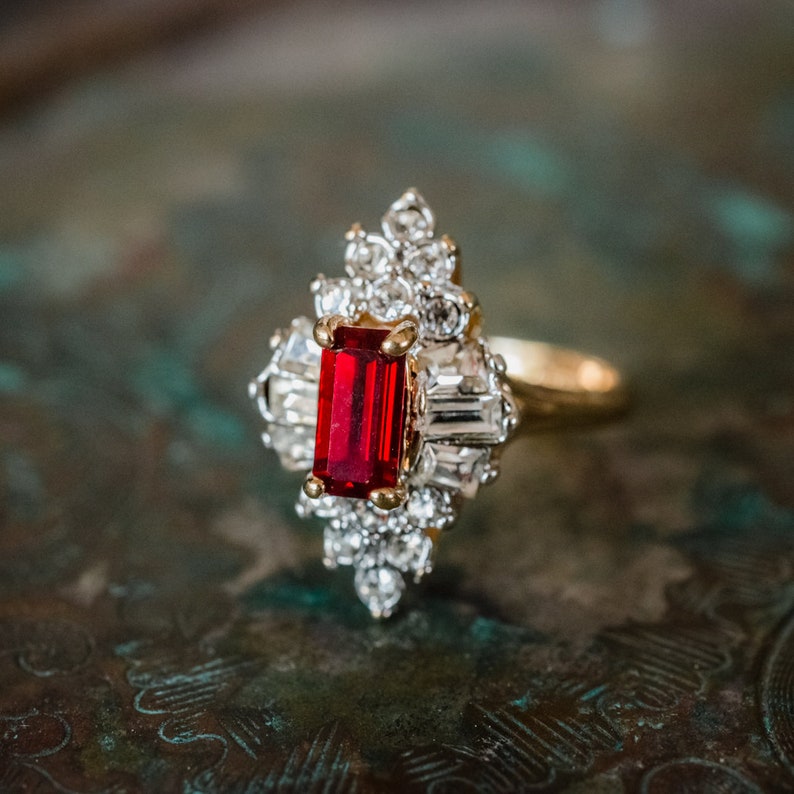 May include: A gold ring with a large red rectangular stone surrounded by smaller clear stones.