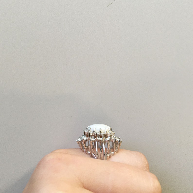 May include: A silver ring with a large white oval stone surrounded by smaller white stones.