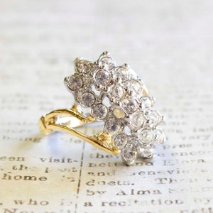 May include: A gold and silver ring with a large cluster of clear rhinestones. The ring is designed in a leaf shape.