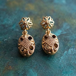 Op de afbeelding: Een paar gouden oorhangers met een bloemmotief. De oorbellen hebben een klein, wit bloemetje bovenaan en een grotere, ovale hanger met een filigraanontwerp.