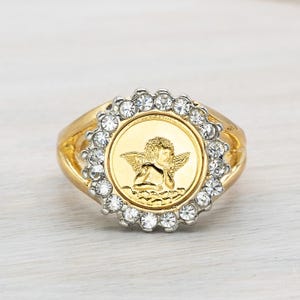 May include: Gold-toned ring featuring a central medallion with an angel design, surrounded by a halo of clear, round gemstones. The ring has a wide band and a polished finish, suitable for everyday wear or special occasions.