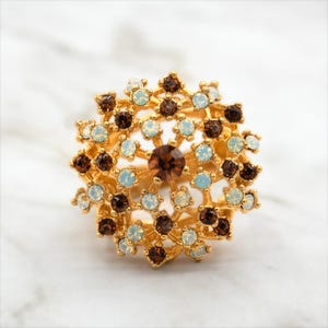 Könnte beinhalten: Ein goldfarbener Ring mit einem großen, komplizierten Design, das aus einer Ansammlung von braunen und weißen Strasssteinen besteht.