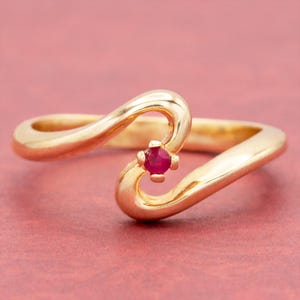 May include: A gold-colored ring with a flowing, asymmetrical design. The ring features a small, round, red gemstone set in the center of the design. The ring is displayed against a pink background.