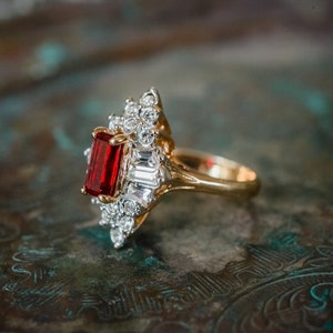 May include: A gold ring with a large red gemstone surrounded by smaller clear gemstones.