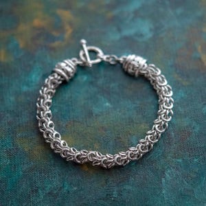 Puede incluir: Una pulsera plateada con cierre de palanca. La pulsera est&aacute; hecha de peque&ntilde;os anillos entrelazados, creando una apariencia texturizada similar a una cota de malla. El fondo es un veteado turquesa y verde.