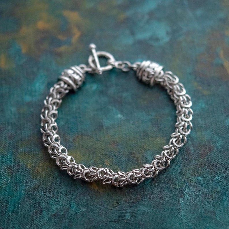 Puede incluir: Una pulsera plateada con cierre de palanca. La pulsera est&aacute; hecha de peque&ntilde;os anillos entrelazados, creando una apariencia texturizada similar a una cota de malla. El fondo es un veteado turquesa y verde.