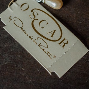 May include: A cream-colored tag with the name "OSCAR" in a gold oval and the words "by Oscar de la Renta" printed in gold. The tag is attached to a jewelry piece with a pearl-like bead.