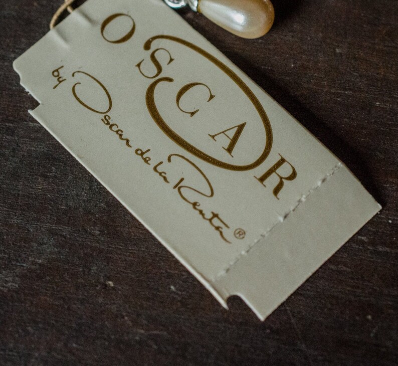 May include: A cream-colored tag with the name "OSCAR" in a gold oval and the words "by Oscar de la Renta" printed in gold. The tag is attached to a jewelry piece with a pearl-like bead.