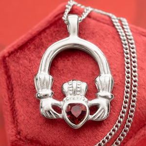 May include: A silver-toned Claddagh pendant necklace with a red heart-shaped gemstone. The pendant features a crown, hands, and a heart, symbolizing love, loyalty, and friendship. The necklace is displayed on a red velvet surface.
