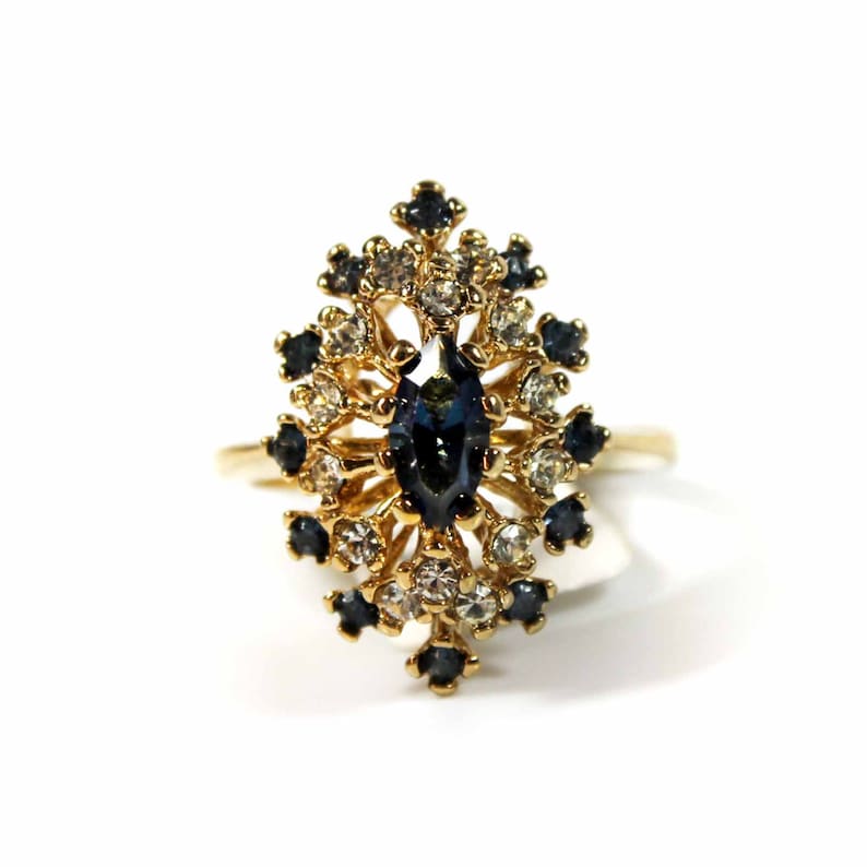 May include: A gold ring with a large blue gemstone surrounded by smaller clear and blue gemstones.