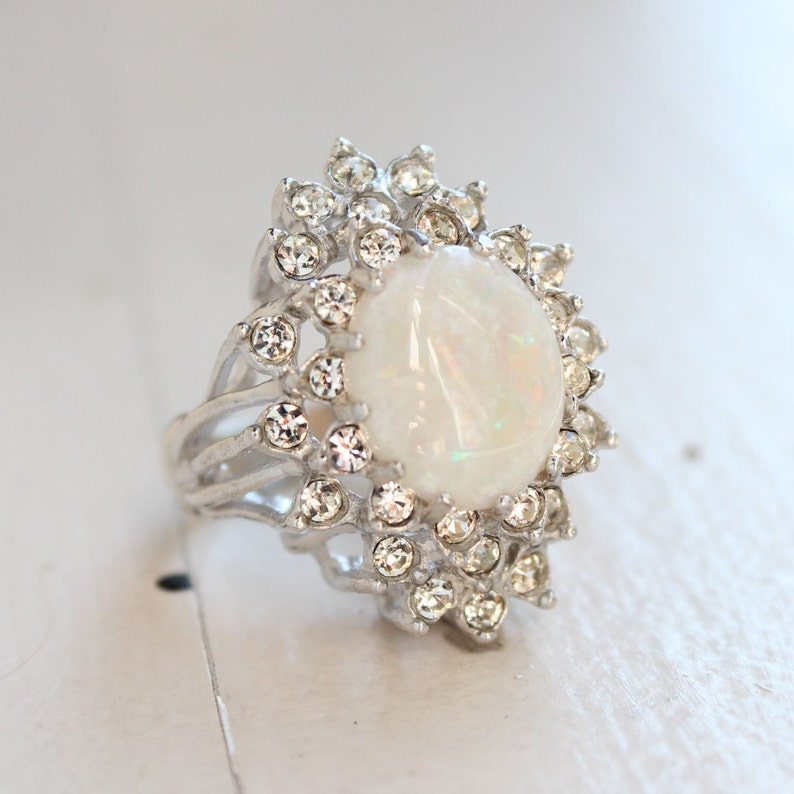 May include: A silver ring with a large white opal stone surrounded by smaller clear rhinestones.