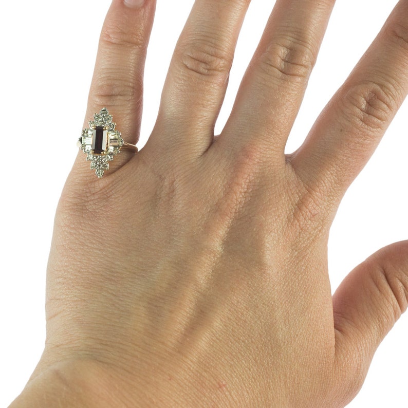 May include: A gold ring with a rectangular dark red gemstone surrounded by small clear gemstones.