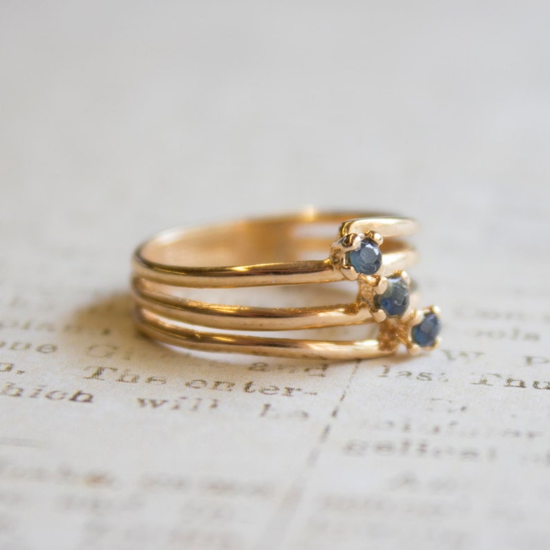 May include: A gold ring with four bands and three blue gemstones. The ring is on a white surface with text.