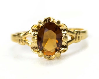 May include: A gold ring featuring an oval, amber-colored gemstone. The gemstone is set in a floral-shaped setting. The band has a textured design. The ring is displayed against a white background.