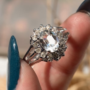 May include: A silver-toned ring featuring a large, oval-cut clear gemstone at the center, surrounded by smaller, round clear stones. The ring has a halo design and a split shank. The ring is held up by a person's fingers.