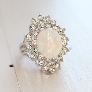 May include: A silver ring with a large white opal stone surrounded by a halo of smaller clear rhinestones.