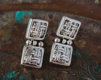 May include: A pair of silver dangle earrings with a woven pattern. The earrings are made of two square pieces connected by a small silver bead.