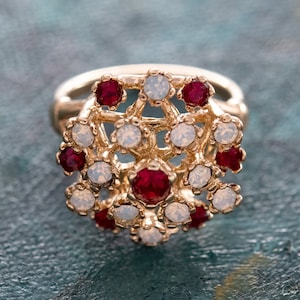 May include: A gold ring with a delicate design featuring red and white gemstones. The ring is set with a cluster of small, round stones in a floral pattern.