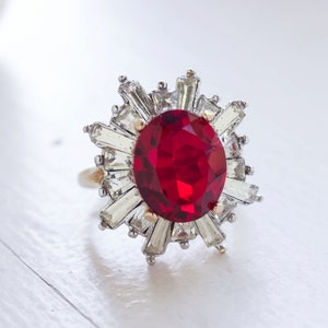 Può includere: Un anello con una grande pietra rossa incastonata in argento e un gruppo di strass trasparenti attorno alla pietra centrale.