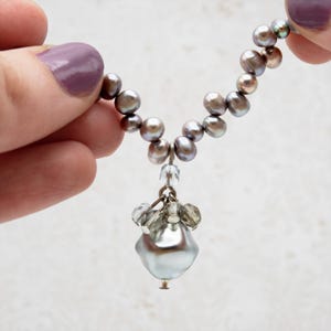 May include: A necklace featuring a strand of gray pearls, with a cluster of clear crystals and a large, pear-shaped, silver-gray pearl pendant. The necklace is held by a hand with purple nail polish.