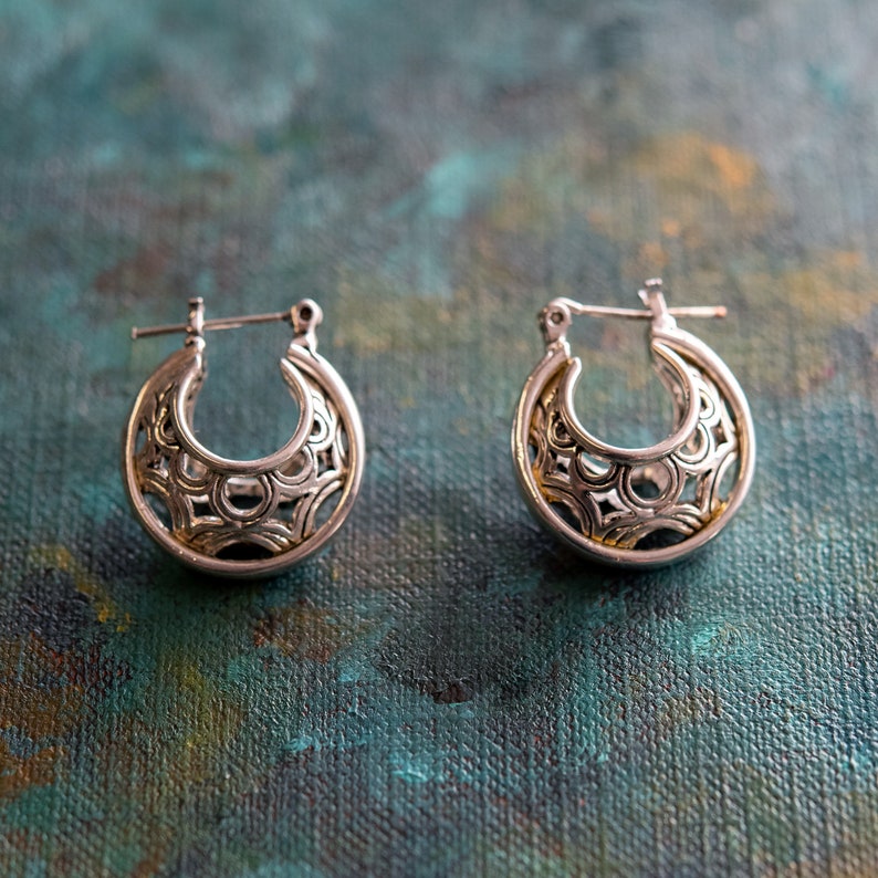 May include: A pair of silver hoop earrings with a delicate, intricate design. The earrings are made of a silver metal with a gold-toned finish.