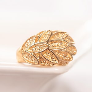 May include: A gold-toned ring featuring a cluster of textured, leaf-shaped elements arranged in a layered, overlapping pattern. The surface of each leaf has a granular, stippled finish, while the edges are smooth and polished, creating a dimensional look.