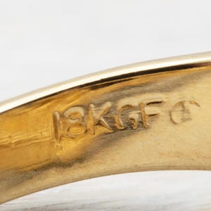 May include: Close-up of a gold ring's interior, showcasing the stamped markings "18K GF". The ring's surface is smooth and reflective, with a warm, golden hue. The markings indicate the gold content and the type of gold used in the ring's construction.