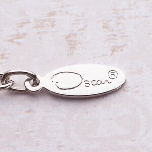 May include: Close-up of a silver oval tag with the name "Oscar" engraved on it. The tag is attached to a silver chain. The background is a light, textured surface.