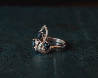 May include: A silver ring with three blue gemstones set in a delicate, intricate design. The ring is on a dark blue surface.