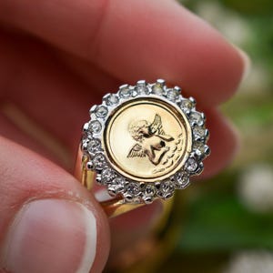 May include: A gold ring featuring a circular design with a cherub image in the center. The cherub is surrounded by a halo of small, clear gemstones. The ring has a gold band.