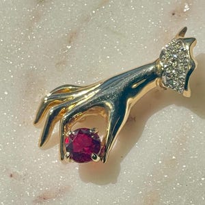 Op de afbeelding: Een goudkleurige broche in de vorm van een hand die een ronde, dieprode edelsteen vasthoudt. De manchet van de hand is bezet met kleine, heldere kristallen. De broche heeft een gepolijste afwerking.