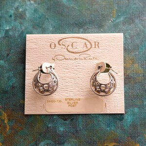 May include: A pair of silver hoop earrings with an intricate design. The earrings are on a white card with the text "OSCAR by Oscar de la Renta" and "STERLING SILVER POST" printed on it. The product number "24465-739" is also printed on the card.