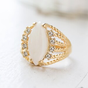 May include: A gold ring with a white oval-shaped stone and clear rhinestones around the stone.